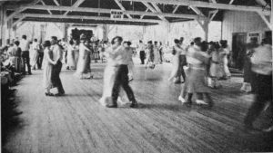 Large dance hall with wooden floors and wooden rafters. Many couples dressed formally are dancing cheek-to-cheek.