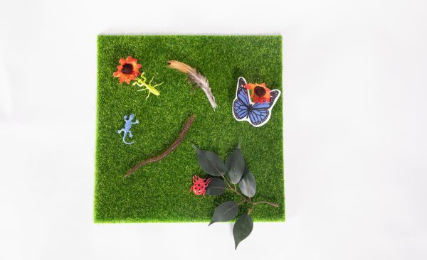 The artificial turf mat serves as the backdrop for a display of several other items from the In My Yard Theme Pack: spray of leaves, matching red flowers, insects, an earthworm, blue butterfly, and bird feather.