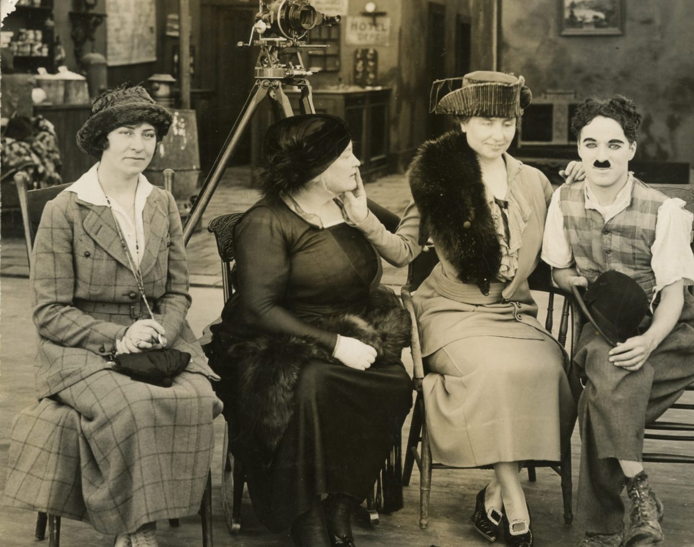 Polly Thomson in a plaid suit and a brimmed hat; Anne Sullivan wearing a black dress and a feathered hat; Helen Keller wearing a light suit, fur stole, and cocked hat; and actor Charlie Chaplain wearing loose pants and a plaid vest. He wears white face makeup and his trademark short mustache and holds a bowler hat in his lap. Keller’s left hand rests easily on Chaplain’s shoulder. Her right hand has two fingers on Sullivan’s lips, and her right thumb rests under Sullivan’s chin. A hand-cranked motion picture camera on a tripod is in the background.