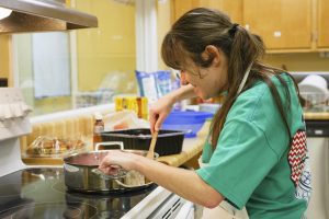 A young woman in a kitchen setting cooking in a sauce pan and a wooden spatula.