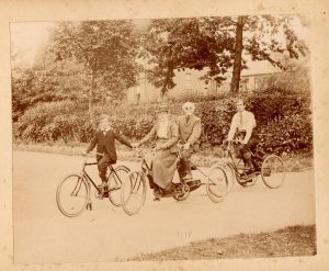 A young boy on a bicycle leads Lady and Sir Francis Campbell, both riding their tandem tricycle, by the hand. Following them on his own adult tricycle is Charles F.F. Campbell.