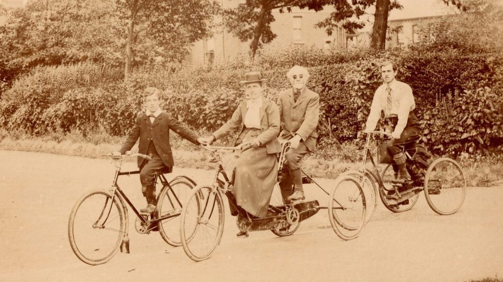 A young boy on a bicycle leads Lady and Sir Francis Campbell, both riding their tandem tricycle, by the hand. Following them on his own adult tricycle is Charles F.F. Campbell.
