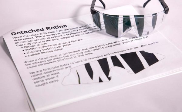 A pair of safety glasses containing the Detached Retina insert sits on top of a printed page with information about the condition and an image of the insert laid flat