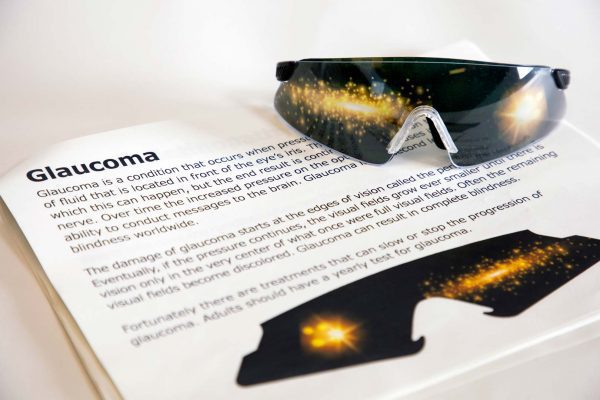 A pair of safety glasses containing the Glaucoma insert sits on top of a printed page with information about the condition and an image of the insert laid flat