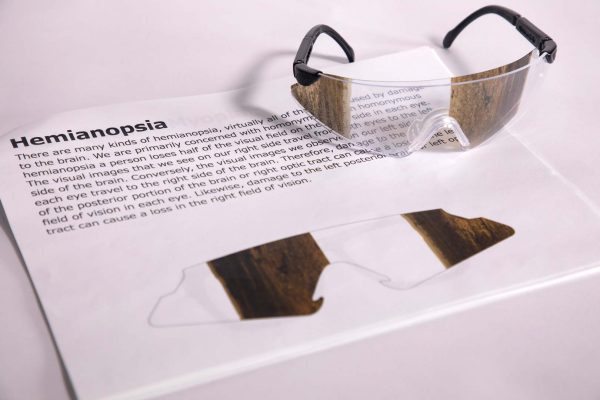 A pair of safety glasses containing the Hemianopsia insert sits on top of a printed page with information about the condition and an image of the insert laid flat.