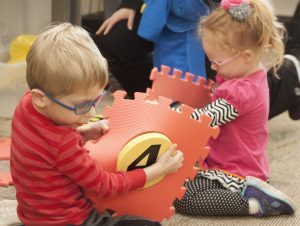 A preschool-aged boy and girl wearing glasses on the floor plaiying with hop-a-dot mats.