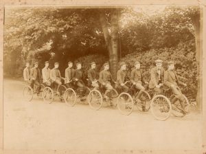 Eleven uniformed students from the Royal Normal College for the Blind pedal between six sets of wheels on a large cycle. An instructor steers from a position second from the front.