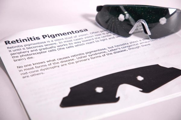 A pair of safety glasses containing the Retinitis Pigmentosa insert sits on top of a printed page with information about the condition and an image of the insert laid flat