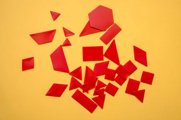 Red tactile manipulatives resting on a yellow background. The red manipulatives include rhombuses, triangles, squares, rectangles, and a hexagon.