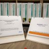 A Brigance supplemental teacher manual binder opened to the cover page and a Brignace braille edition assessment binder opened to the cover page sit on top of a wooden table with additional Brigance binders shown on a bookshelf in the background.