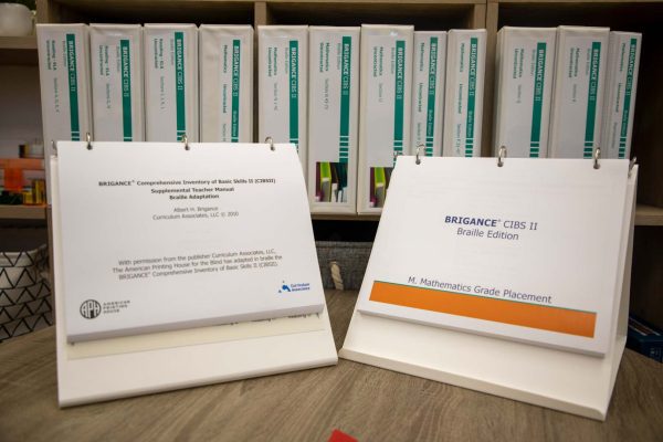 A Brigance supplemental teacher manual binder opened to the cover page and a Brignace braille edition assessment binder opened to the cover page sit on top of a wooden table with additional Brigance binders shown on a bookshelf in the background.