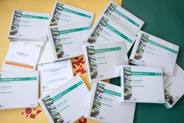 Multiple uncontracted braille Brigance binders spread across a yellow and green surface. Red tactile shapes like a hexagon, rhombus, and triangle can also be seen spread across the table.