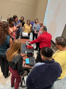 A group of 11 people standing around a table looking and touching different tech including PageBlaster, Mantis Q40, and Chameleon 20.