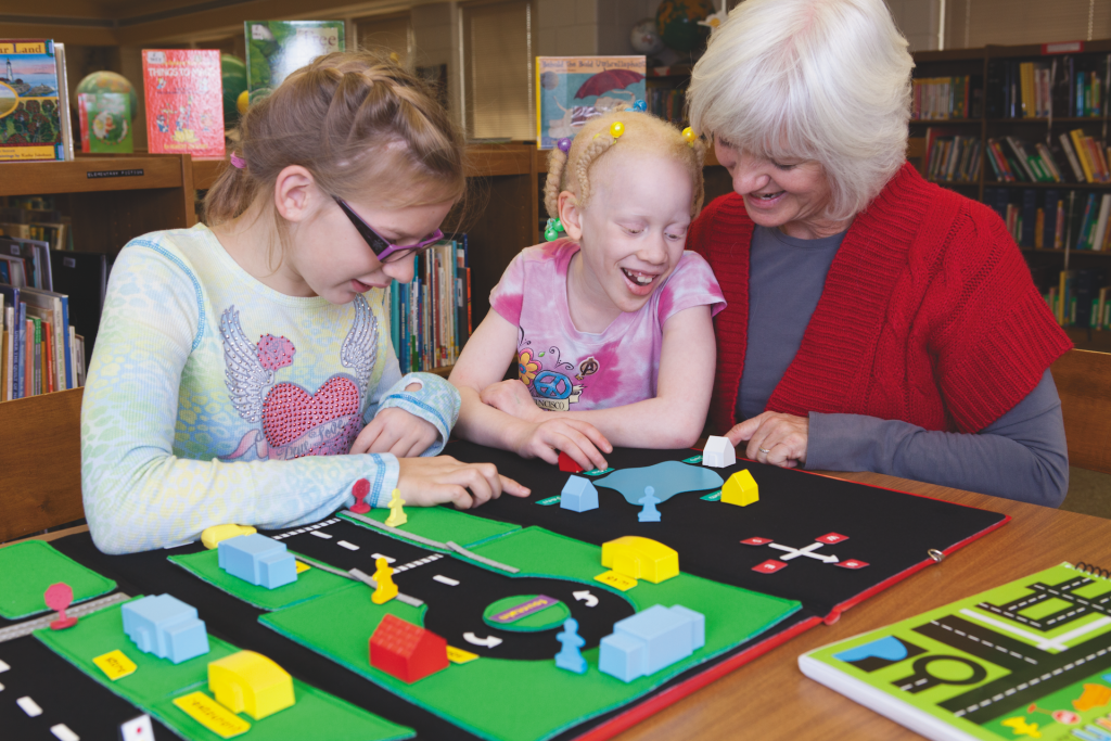 Two girls and a woman in a library setting play with Tactile Town. Tactile Town is made up of a black board with pieces that represent, grass, street crossings, and different kinds of buildings in bright colors like green, red, blue, and yellow.