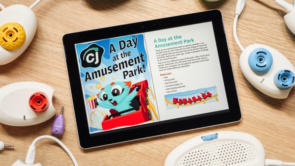 A tablet sitting on a table displaying the Code Jumper Puzzle – A Day at the Amusement Park. On the tablet’s screen is an image of CJ, a blue cat-like creature riding a roller coaster with a box of popcorn in one and hand the other hand up in the air. Code Jumper pods lay on the table surrounding the tablet.
