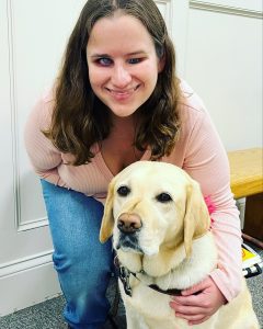 Jess, wearing jeans and a light pink shirt, smiles at the camera. Her arm is around her guide dog, Joyce, a yellow lab wearing a harness.