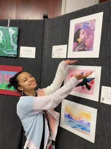Young girl poses with her hands directly below the artwork she created. Her piece is a self-portrait featuring her looking towards the sky with a pleased expression and purple, pink, blue and dark colored paint on her face and in the background.