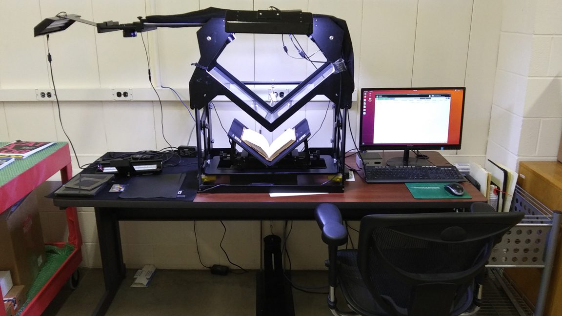 A large scanner, 2’ wide x 2 1/2’ tall, resting on a tabletop, hooked up to a computer. Two v-shaped “cradles,” one big, made of glass, and another half its size, made of metal, cross the window from side to side. A book lies open on the smaller cradle.