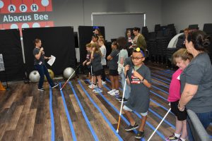 A group of students and a few adults, many holding white canes and wearing APH Abacus Bee shirts. Tactile tape stripes the floor and the APH Abacus Bee logo is projected on the back wall.