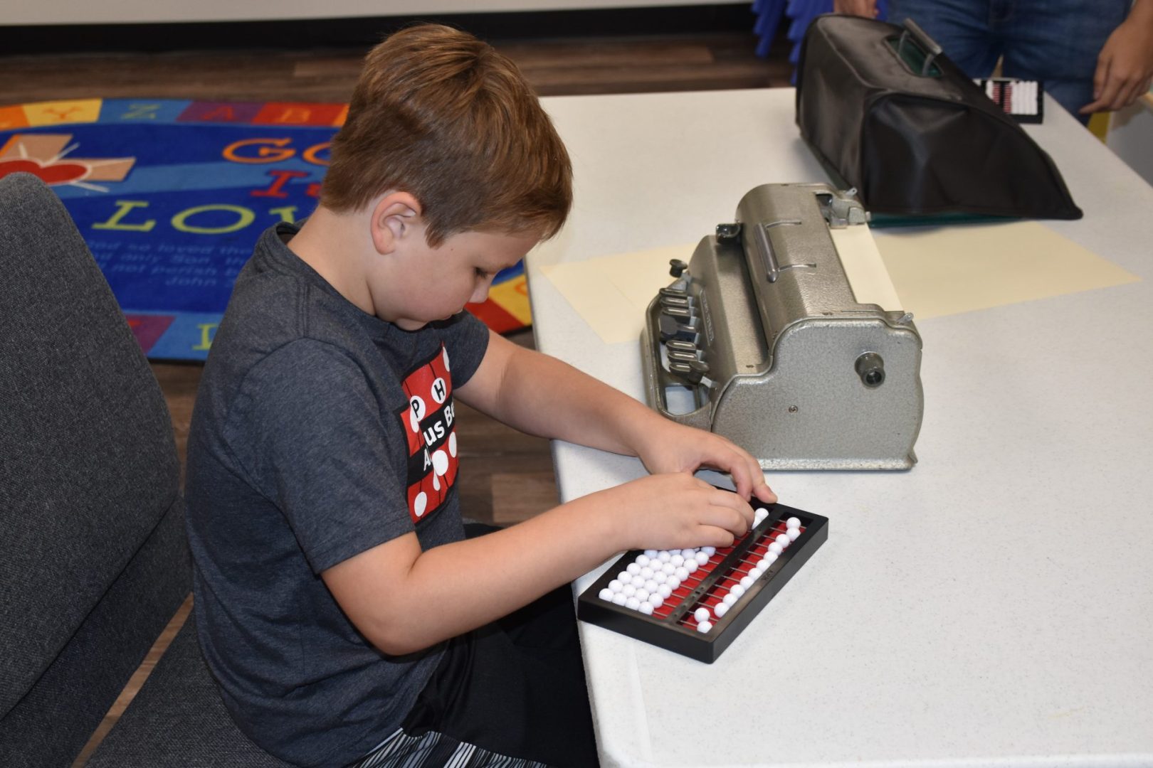 A boy in an Abacus Bee t-shirt sits at a table using an abacus with a braillewriter to the side.