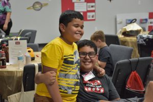 Two boys smile with one arm around each other. One boy has glasses and is wearing an APH Abacus Bee shirt.