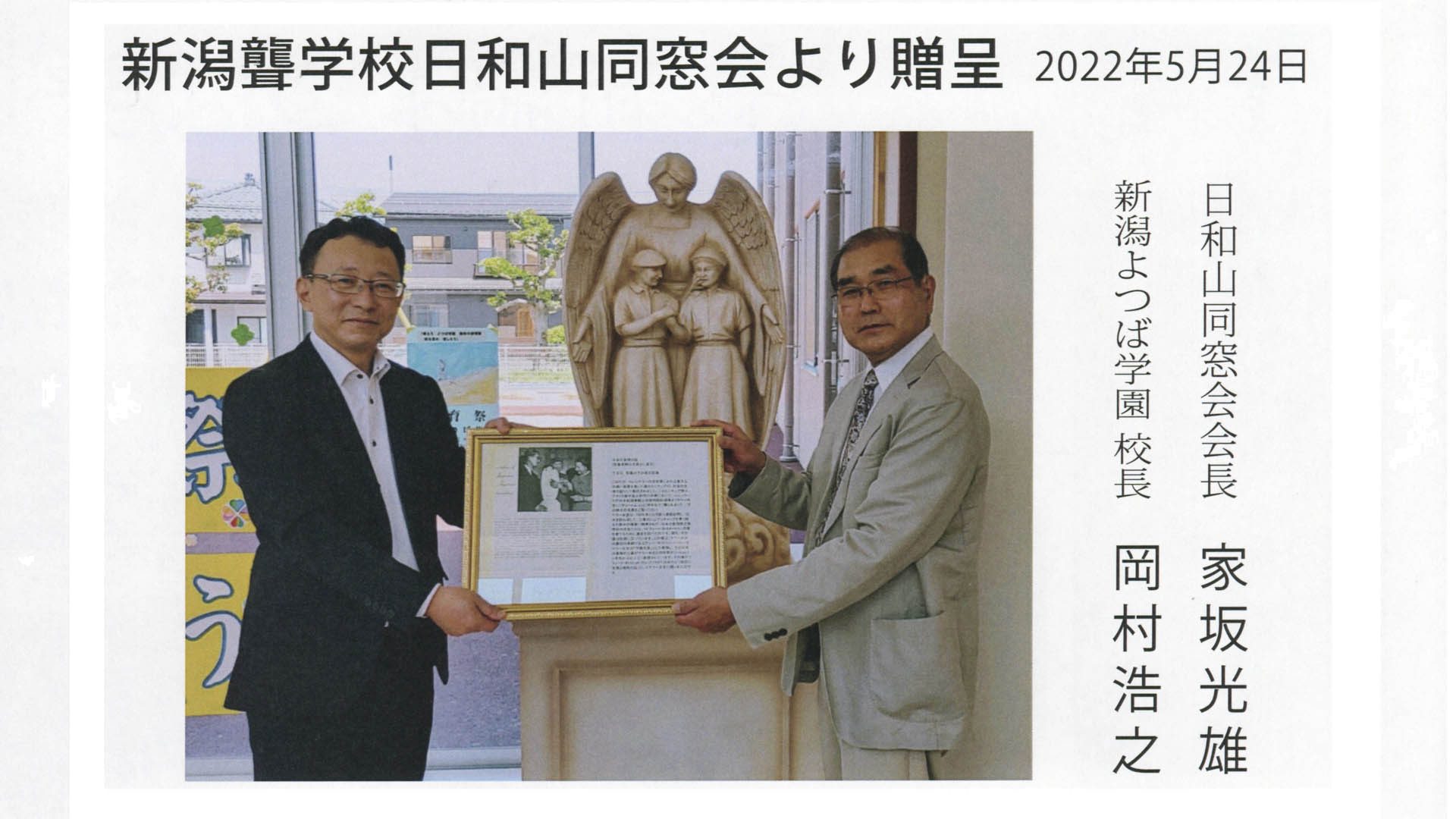 Two men stand in front of a 24” statue of Anne Sullivan, with the wings of an angel, embracing Helen Keller and Polly Thompson as they use the manual alphabet. The men hold a framed article in Japanese showing a similar statue being presented to Helen Keller. The photo is surrounded by text in the Japanese language.