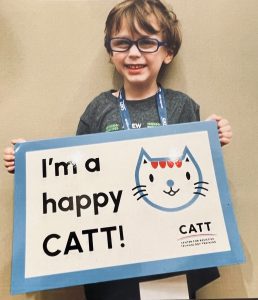 A smiling young boy with glasses holding a "I'm a happy CATT" sign with a kitty logo.