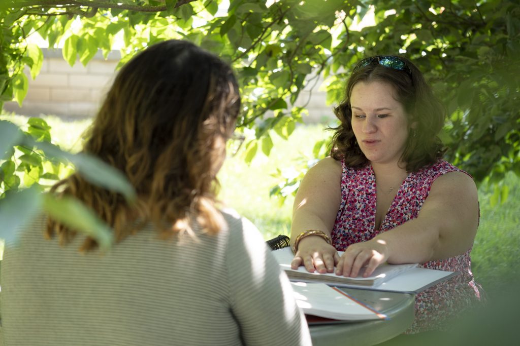 Two college-aged women sit at a table under a tree reading textbooks.