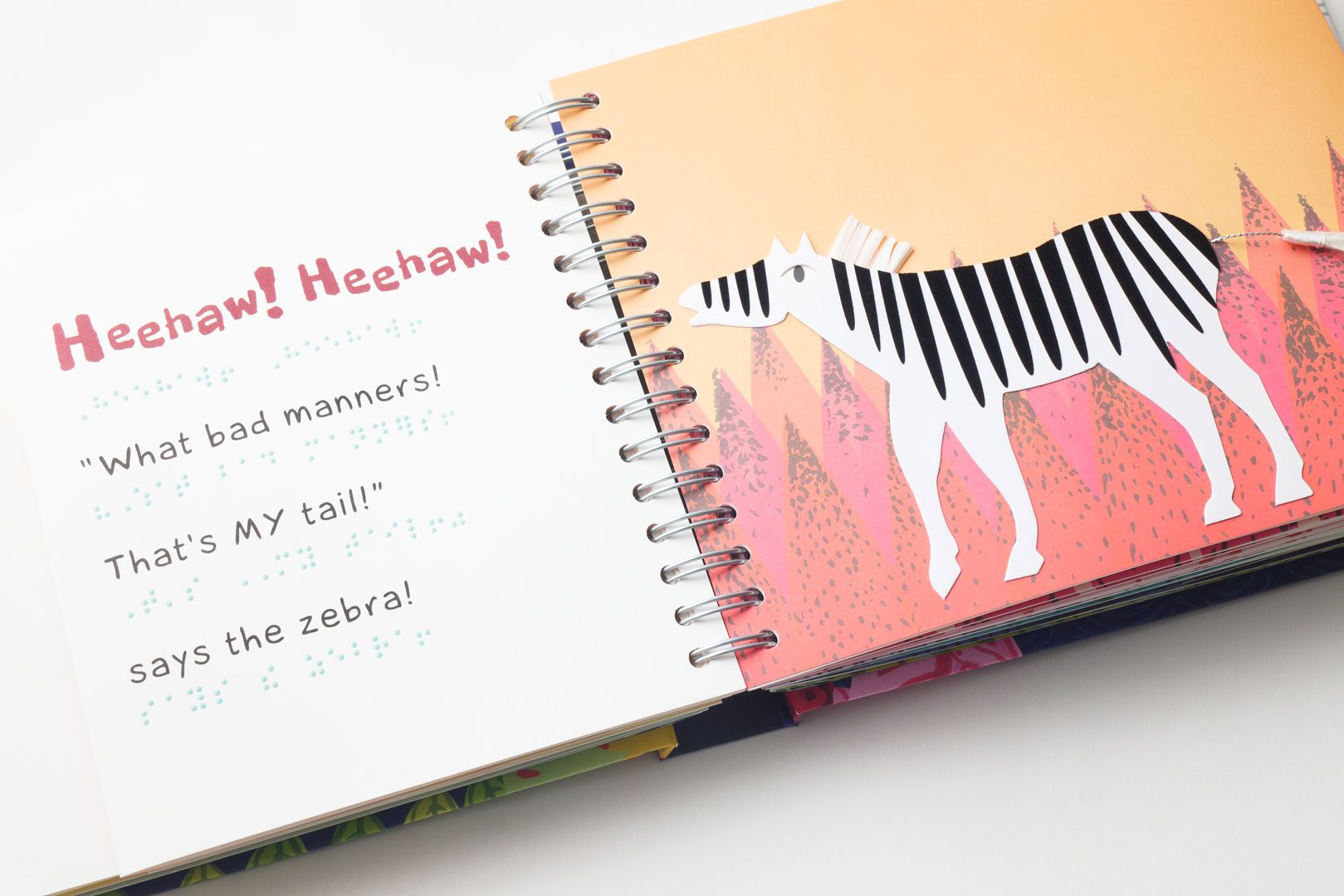 The book is opened to show two pages. On the left-hand page in large pink text reads, “Heehaw! Heehaw!.” Underneath that in large text reads, ““What bad manners! That’s MY tail!” says the zebra!” Under each line of large print text is the braille translation. On the right-hand page is a white and black striped zebra on top of a peach and pink background. The zebra’s tail is made from rope and a small white brush.