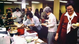 A photo from the 70s of a potluck table with a Christmas tree in office area with office staff lined up and filling plates.
