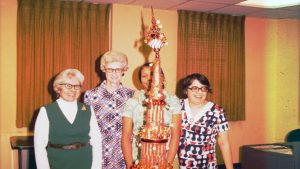 A photo from the 70s of four ladies stand side by side at a table decorated with a small red sleigh and a towering Christmas 