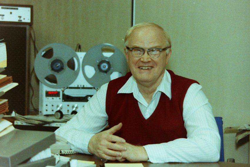 Raymond Randles, a white man with a high forehead and sandy blonde hair wearing thick eyeglasses, a red sweater, and a white collared short, sitting at a desk with a reel-to-reel tape machine in the background. He grins at the photographer.