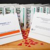 A Brigance supplemental teacher manual binder opened to the cover page and a Brigance braille edition assessment binder opened to the cover page sit on top of a wooden table with additional Brigance binders shown on a bookshelf in the background. Red manipulatives sit on the table between the two binders.