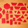 Red tactile manipulatives resting on top of a wooden surface. The red manipulatives include rhombuses, triangles, squares, rectangles, trapezoids, and a hexagon.