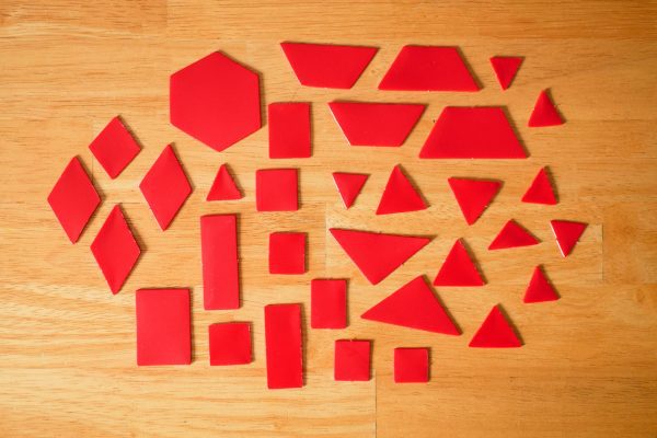 Red tactile manipulatives resting on top of a wooden surface. The red manipulatives include rhombuses, triangles, squares, rectangles, trapezoids, and a hexagon.