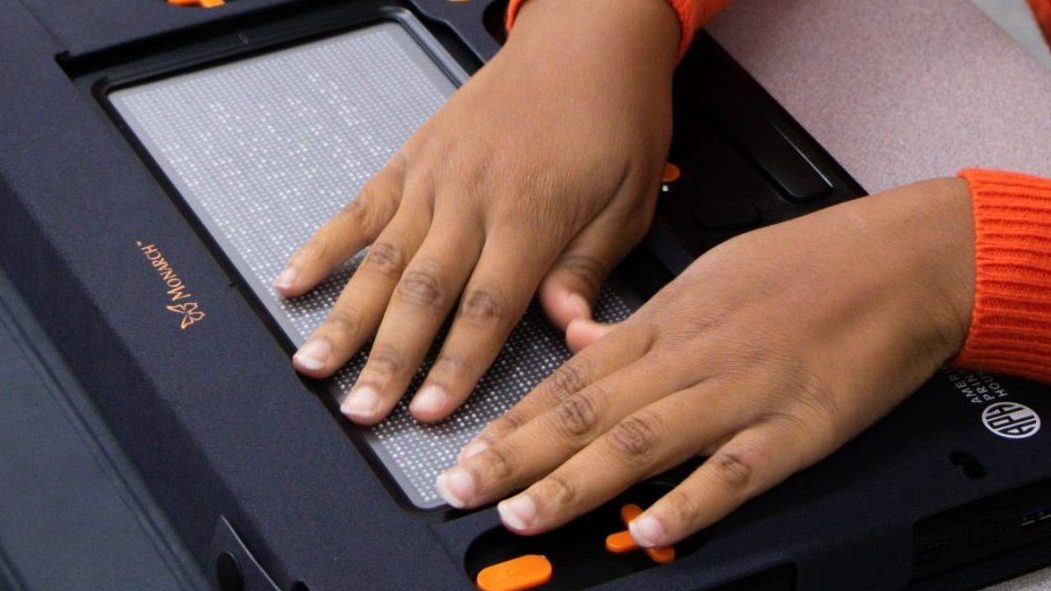 Hands on braille being displayed on a 10 line by 32 cell refreshable device with orange buttons.
