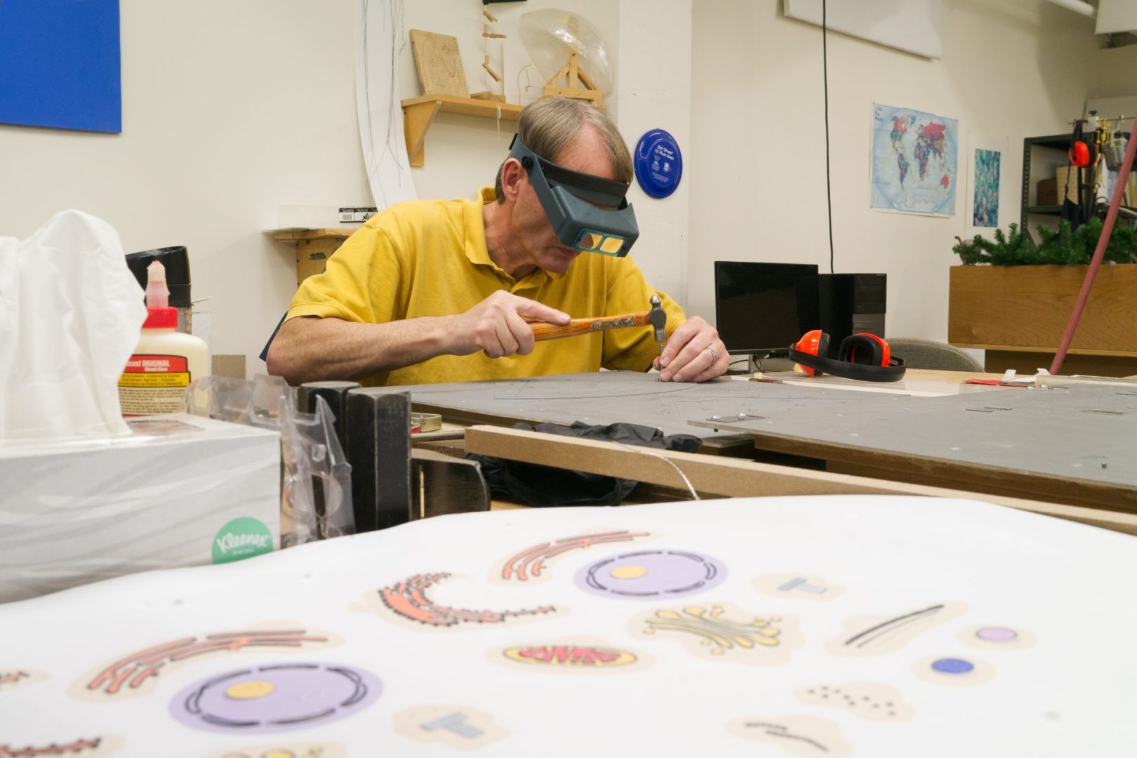 A man leans over a worktable, using a hammer. He is wearing a magnifier on his head.