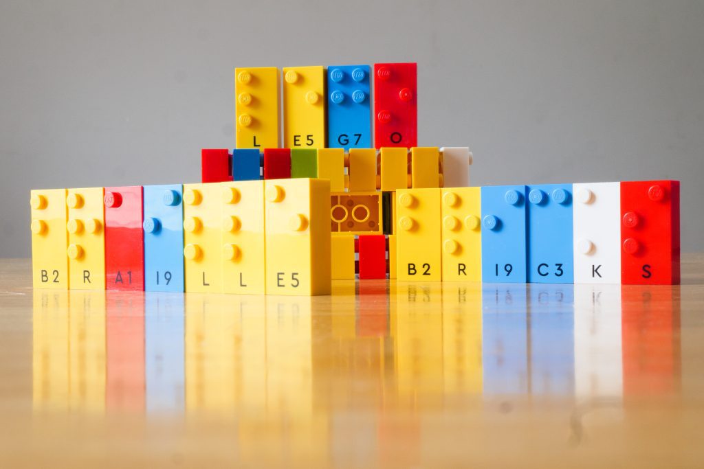LEGO Braille Bricks standing up on a table. They are in a variety of primary colors and feature studs in a variety of braille cell configurations. Some of the bricks are lined up to spell 