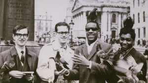 Black and white snapshot, left to right, unknown man, Ted Hull, Stevie Wonder, and Hamilton Bohannon in Trafalgar Square, London on concert tour; Wonder and Bohannon have pigeons standing on their heads and arms; a base for a statue and a church are in the background; Hull is wearing a light trench coat, Wonder is smiling and wearing dark sunglasses and a wool coat, Bohannon is smiling at the camera and wears a dark jacket.