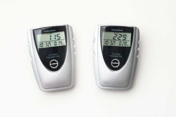 Two, white and black, walking pedometers on a white background. The pedometer on the left shows 115 steps, 0.14 miles walked, and a duration of 10 minutes and 32 seconds. The pedometer on the right, shows 229 steps, 0.28 miles walked, and a duration of 10 minutes and 32 seconds.