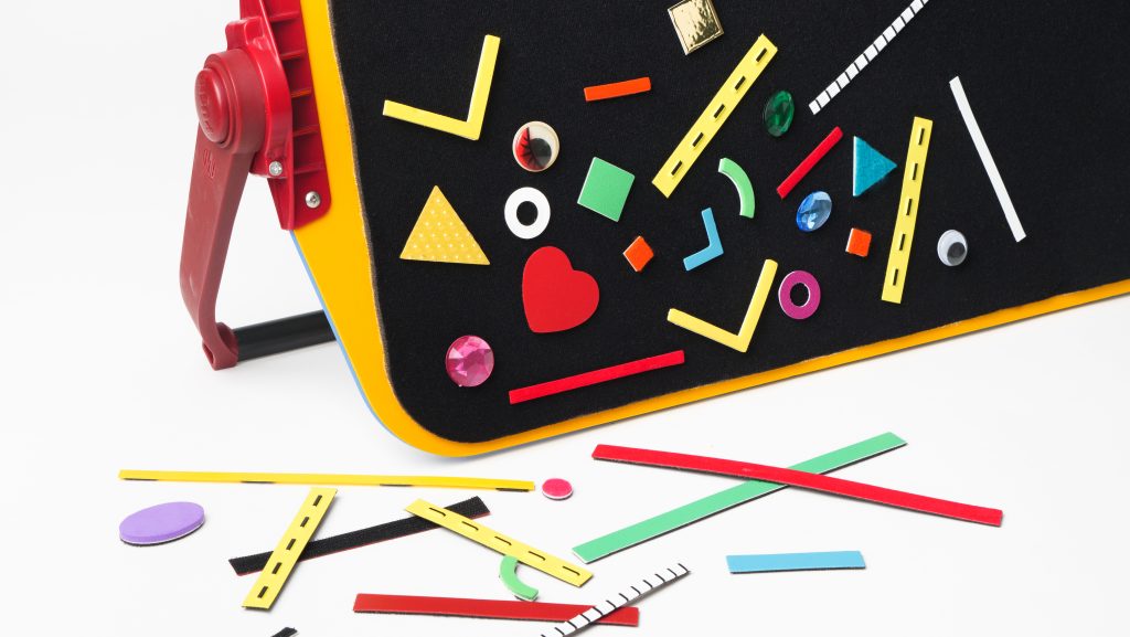 The corner of the velcro side of an All-in-One Board decorated with an array of colorful tactile shapes. There is also an assortment of tactile shapes scattered in front of the All-in-One Board.