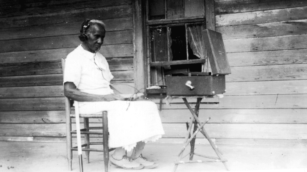 An old woman sits on straight-backed chair on a porch. Her cane rests against the chair. She wears headphones, with wires that lead to a talking book player set on a nearby table.