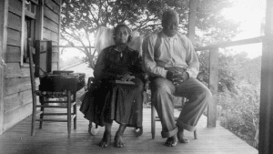 A barefoot couple sit on their porch, with the country wilderness in the background. The woman wears earphones and listens to a talking book player. The man sits quietly beside her, his hands folded in his lap.