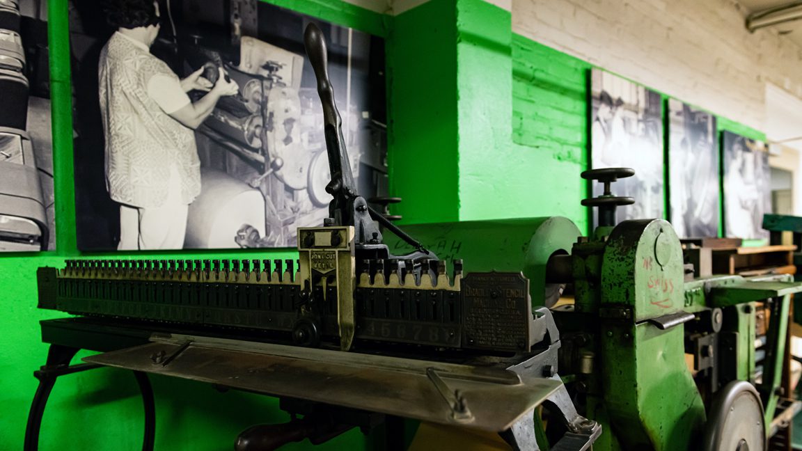 A view of one wall of the Museum Annex. Visible are two industrial machines. One resembles a piano, except each key makes a punch onto a piece of metal. The second shows three rounded tops (part of a cylinder press). Blurred images of other machines continue down the wall.