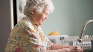 An elderly woman washes her hands at a kitchen sink.