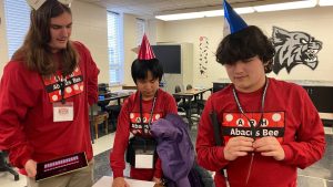 Three students wearing shirts with the Abacus Bee logo and party hats stand around a table. The student in the middle is reading a brailled piece of paper.
