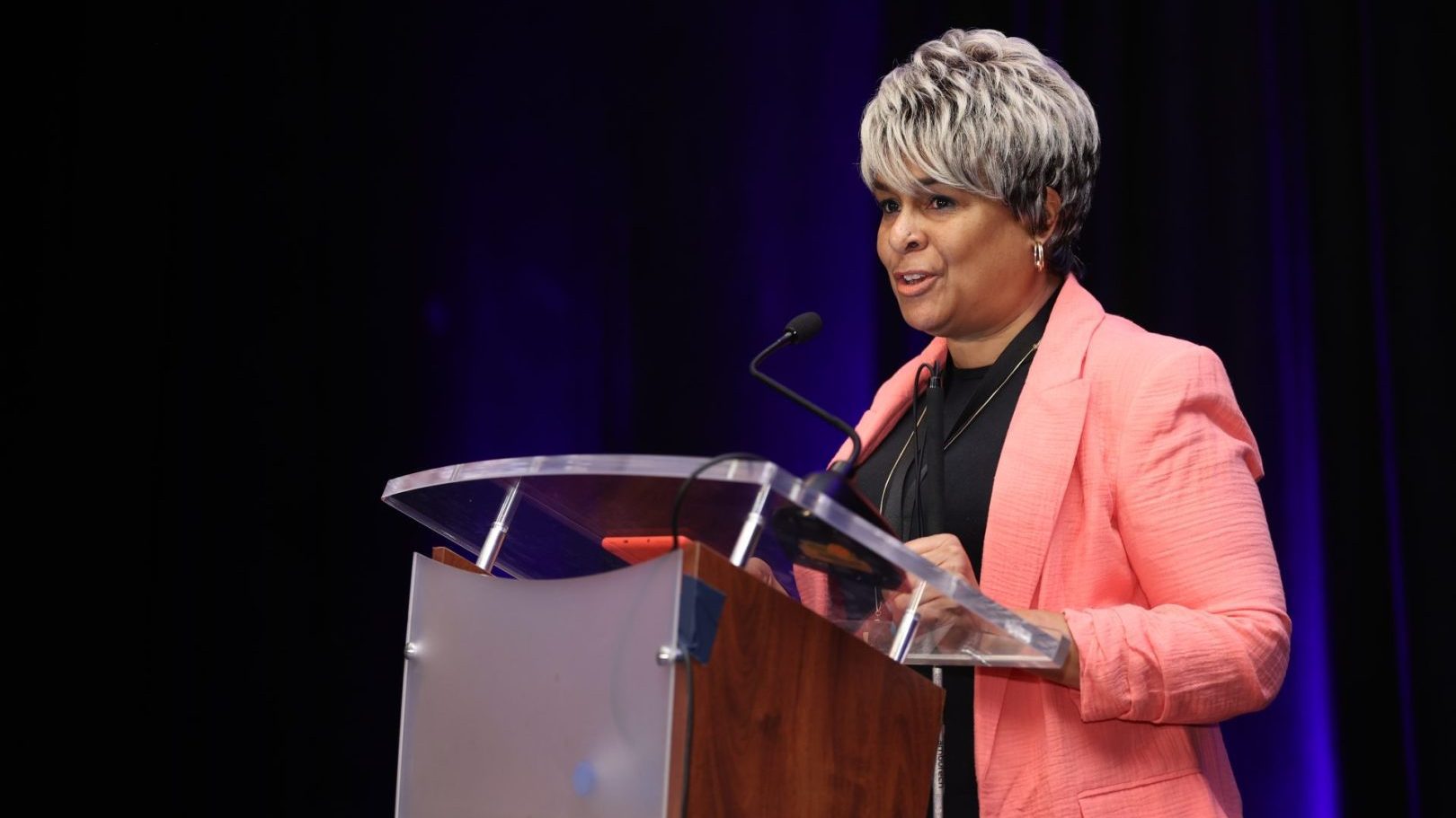 A woman wearing a coral pink jacket speaks from a podium.