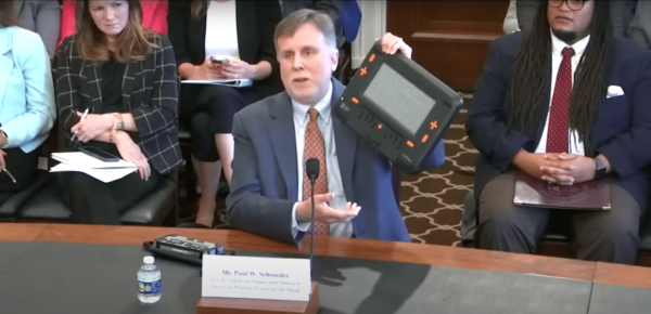 In a screengrab of a livestream video, Paul Schroeder holds up the Monarch, a multiline refreshable braille device with orange buttons and an array of refreshable braille pins, while speaking into a microphone at the front of a seated crowd of people.