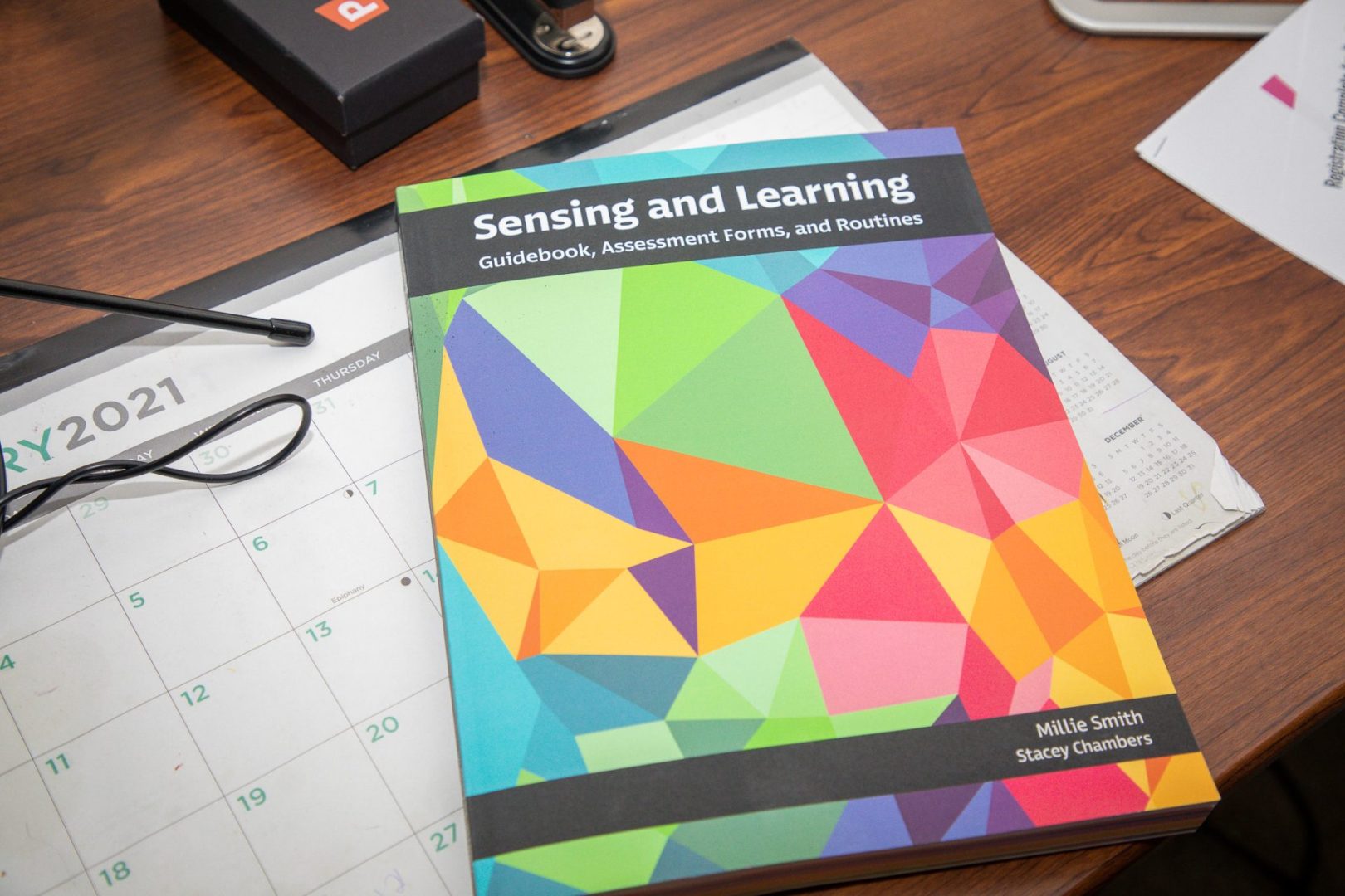 A copy of Sensing and Learning lays on a wooden desk on top of a desk calendar and beside some office supplies.