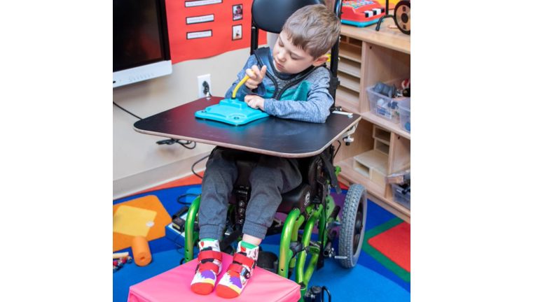 A young learner sitting in a wheelchair with his feet resting on APH’s Vibrating Pad, uses his hands to activate the vibration using APH’s Adaptable Stick Switch, which sits on the wheelchair tray.